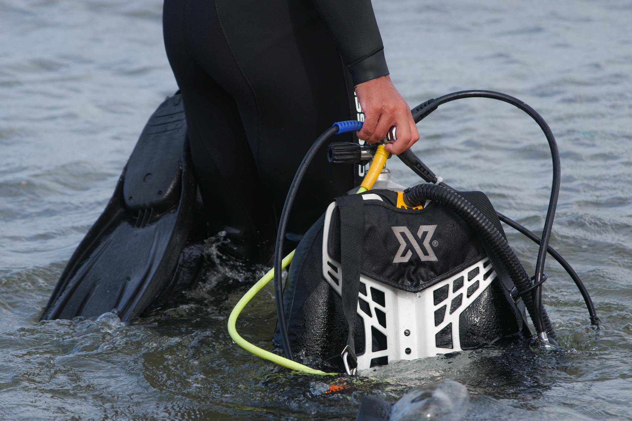 A diver in a wetsuit carrying scuba equipment while wading through water.