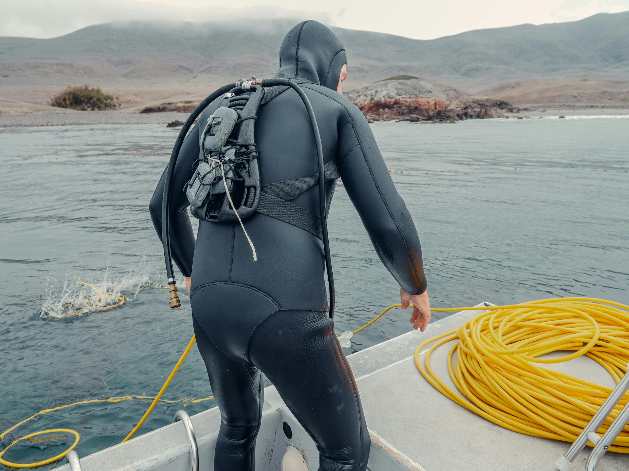 Scuba diver in wetsuit ready to explore the ocean from a boat, near rocky shore.